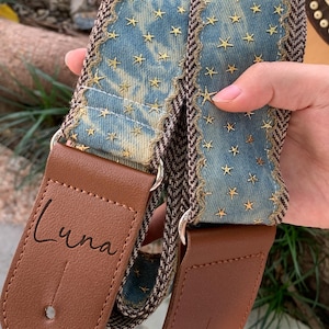 May include: A guitar strap featuring a denim blue central section decorated with gold star embroidery. Brown leather ends are present, with the word "Luna" printed on one. The edges are trimmed with a brown and cream herringbone pattern.