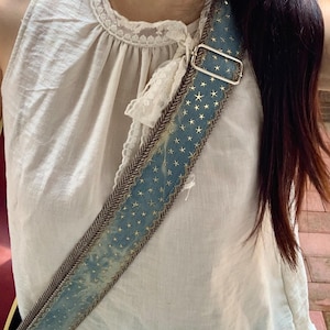 May include: A blue and gold star-patterned guitar strap with a brown herringbone border and a silver metal buckle. The strap is worn over a white top with lace detailing.