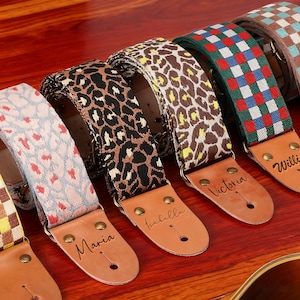May include: A selection of guitar straps with diverse patterns, such as floral, leopard print, and checkered designs. Each strap has a leather end with a name engraved. The straps are presented on a wooden surface.