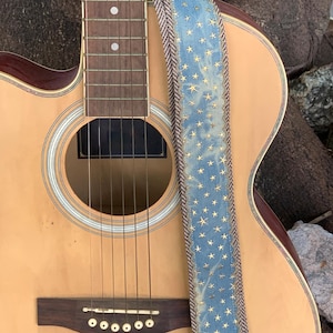 May include: Acoustic guitar with a light brown body and a blue guitar strap. The strap is decorated with gold stars and a brown and white patterned border. The guitar has six strings and a dark brown bridge.