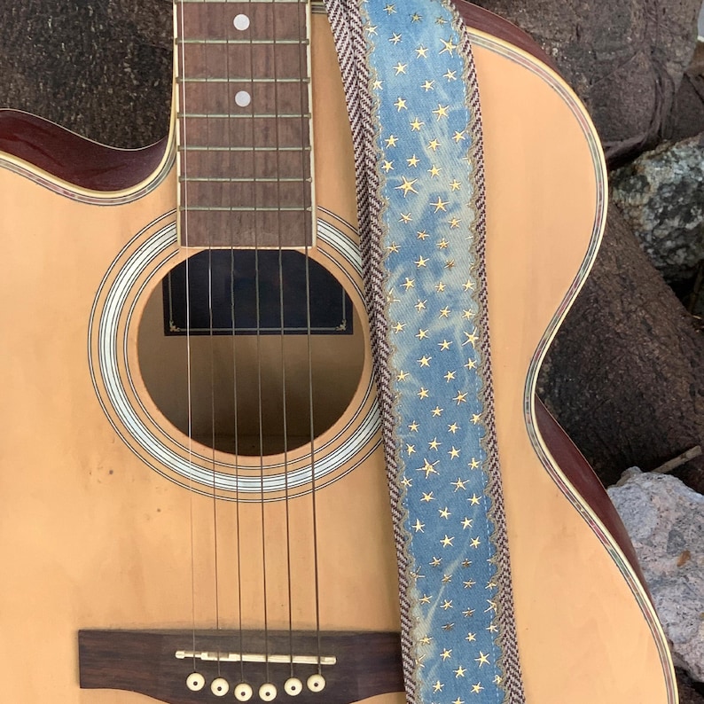 May include: Acoustic guitar with a light brown body and a blue guitar strap. The strap is decorated with gold stars and a brown and white patterned border. The guitar has six strings and a dark brown bridge.
