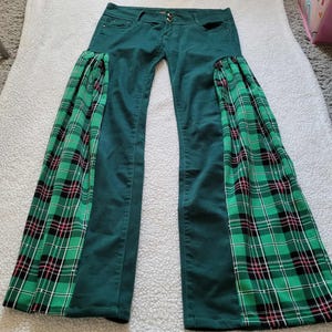Puede incluir: Pantalones vaqueros verdes con paneles acampanados de tela a cuadros verdes, negros y rojos. Los pantalones tienen cierre de botón y cremallera y están colocados sobre una superficie blanca.