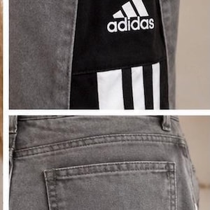 May include: Grey denim trousers featuring a high-waisted design with a button closure. The sides display a black panel with three white vertical stripes and a white Adidas logo printed on the upper section. The trousers include a back pocket.