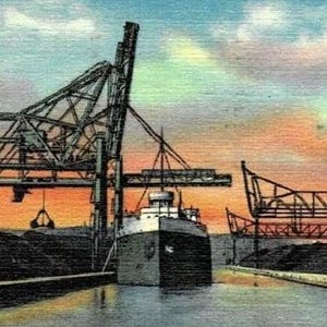 May include: Vintage postcard featuring the 133-D Coal Docks in Duluth-Superior Harbor. The image shows a ship navigating a waterway, framed by industrial structures and coal piles. The sky displays a colorful sunset.