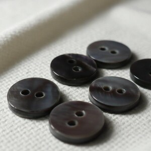 May include: A collection of six round, dark brown buttons, each featuring two central holes for attachment. The buttons have a smooth, slightly iridescent surface with subtle variations in color and a polished finish, arranged in a cluster.