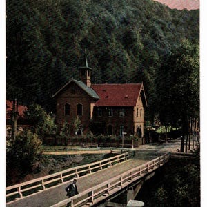 May include: Vintage postcard featuring a church in Treseburg, Germany. The image shows a bridge over a river, leading to a brick building with a red tile roof. Lush green trees cover the hillside in the background.