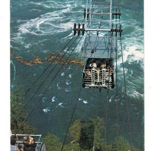 May include: A vintage aerial cable car suspended by thick black cables over a turbulent, deep blue river with swirling white water currents. The metal car features a skeletal frame structure and is positioned above a lush green forest canopy.