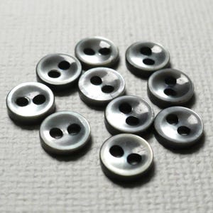 May include: A collection of ten round, dark gray buttons, each featuring a smooth, reflective surface and two centered holes for attachment. The buttons have a slightly iridescent sheen and a uniform circular shape, arranged in a loose cluster.