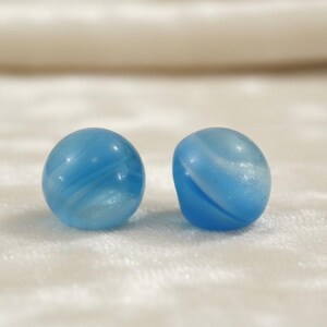 May include: Two round, light blue glass beads featuring a smooth, polished surface with subtle, swirling white internal patterns. These spherical beads exhibit a translucent quality and a consistent, rounded shape, showcasing a soft, glossy finish.