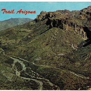 May include: A vintage postcard featuring a scenic view of the Apache Trail in Arizona. The image showcases a winding road through a rugged, mountainous landscape with lush green vegetation and a clear blue sky. The words "Apache Trail, Arizona" are printed in red.