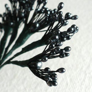 May include: A cluster of artificial floral stamens featuring numerous small, glossy black, teardrop-shaped heads attached to thin, dark green wire stems. The individual stems are bundled together, creating a branched, spray-like arrangement.