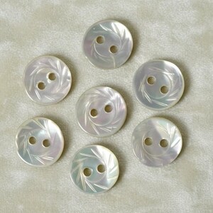 May include: A set of seven round, off-white mother-of-pearl buttons, each featuring two center holes and a decorative engraved pattern of radiating lines along the outer edge, displaying a smooth, iridescent, and glossy surface finish.