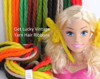 May include: A doll head with blonde hair styled in pigtails tied with bright yellow yarn ribbons. Beside it is a bundle of multi-colored yarn in yellow, orange, green, brown, and cream, with a label reading Get Lucky Vintage Yarn Hair Ribbons.