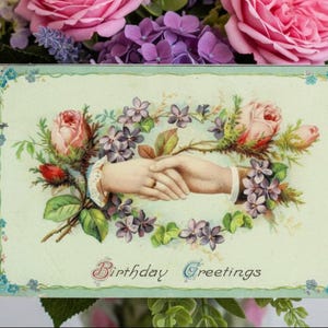 May include: Vintage birthday greeting card with a floral design. The card features two hands clasped together, surrounded by roses and other flowers. The words "Birthday Greetings" are written on the card.