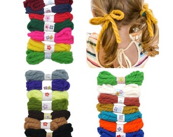 May include: A collection of thick, twisted yarn hair ties in various solid colors and one striped pattern, each secured with a white paper band featuring a small flower icon. The yarn ties are shown bundled and tied around pigtails.