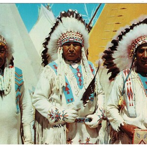 May include: Vintage postcard featuring three Native American men in traditional ceremonial attire. They wear elaborate feathered headdresses, white shirts with turquoise and red accents, and beaded necklaces. The background includes teepees.