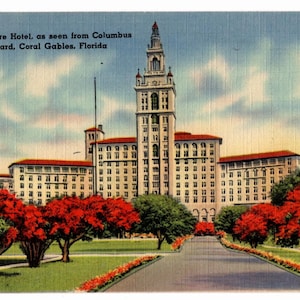 May include: Vintage postcard of the Miami Biltmore Hotel in Coral Gables, Florida. The image shows the hotel's exterior with a tall tower, red-tiled roofs, and a long driveway lined with trees and flowers. The sky is blue with some clouds.