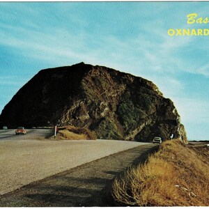 May include: A vintage postcard showing a large, dark brown rock formation known as Bass Rock on the Oxnard Coast. A paved road curves around the base of the rock with two small cars visible. The sky is a clear blue, and the landscape is dry.