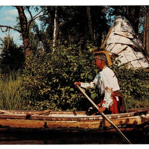 May include: A vintage postcard depicts a Native American man paddling a wooden canoe on a lake. He wears traditional attire, including a headdress. A teepee is visible in the background, surrounded by trees and lush greenery. The scene evokes a sense of history and culture.