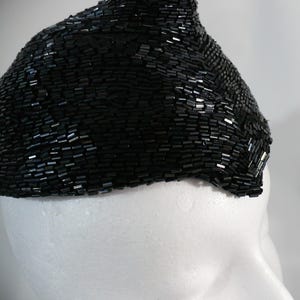 May include: A black beaded head covering, possibly a hat or headband, with a dense, textured surface. The beads are small and rectangular, creating a shimmering effect. The head covering is displayed on a white mannequin head.