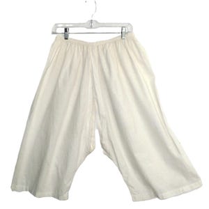 May include: Off-white, wide-leg pants hanging on a clear plastic hanger. The pants have a loose, flowing design, with a gathered waistband. The fabric appears to be a lightweight material, suitable for warm weather.