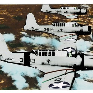 May include: Vintage photograph of three U.S. Navy aircraft in flight. The planes are light gray with black accents and white markings, including the text "U.S. NAVY" and the numbers "3-13" and "3-B-14". The background shows a cloudy sky and land.