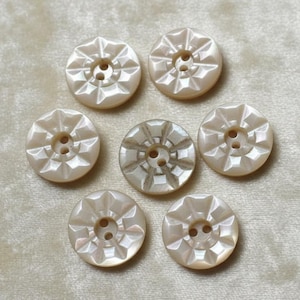 May include: Seven round, off-white buttons with a pearlescent finish. Each button features a geometric, star-like faceted pattern radiating from the center, where two small holes are positioned for attachment to fabric or other materials.