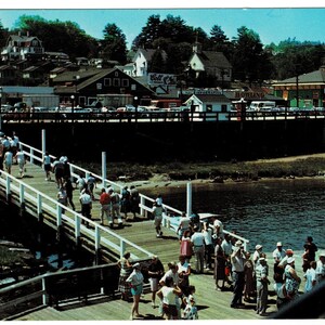 May include: Vintage postcard depicting a bustling pier scene. People stroll along a wooden pier with white railings, leading to a town with buildings and a church. A boat sails on the water. The sky is blue.
