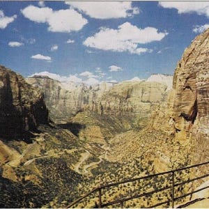 May include: A vintage postcard featuring a scenic view of a canyon with towering rock formations. The image showcases a winding road, lush greenery, and a bright blue sky dotted with fluffy white clouds. A metal railing is in the foreground.