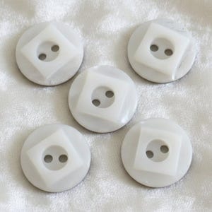May include: Five off-white buttons with a circular shape and a square inset. Each button has two holes for sewing. The buttons are arranged on a white, textured surface.