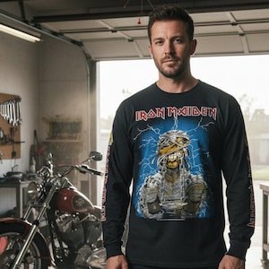 May include: Black long-sleeve shirt featuring the band name "Iron Maiden" above a graphic of a mummy with blue lightning bolts on a blue background. The sleeves have additional text. A motorcycle is visible in the background.