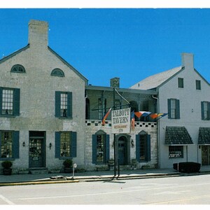 May include: Vintage postcard featuring the Talbott Tavern Restaurant. The building is a two-story structure with a light-colored stone facade, blue shutters, and a sign that reads "Talbott Tavern Restaurant". The sky is a clear blue.