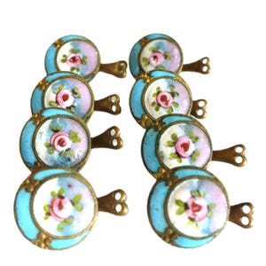 May include: Eight antique drawer pulls with a floral design. Each pull features a round, light blue enamel face with a pink rose and green leaves. The pulls have a brass handle and a decorative brass frame.