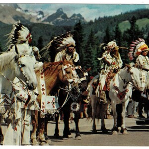 May include: Vintage postcard featuring Native American riders on horseback, adorned in traditional headdresses and attire. The horses are decorated with ceremonial blankets. The background shows mountains and trees, creating a scenic backdrop.