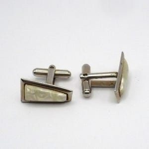 May include: A pair of vintage cufflinks. Each cufflink features a silver-toned metal frame with a cream-colored, textured inlay. The cufflinks have a classic T-bar closure for securing to a shirt cuff.