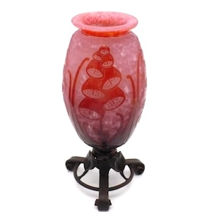 May include: A pink and red glass vase with a mottled texture and etched floral patterns, featuring a wide rim and a tapered body. The vase is supported by a dark metal stand with three curved legs and leaf-shaped accents at the base.