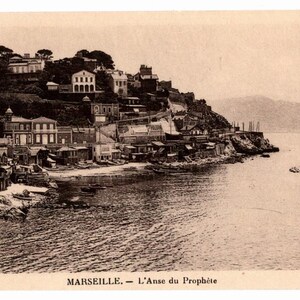 May include: Vintage sepia-toned postcard featuring a coastal scene of Marseille, France. The image shows buildings along the shore and on a hillside, with boats in the water and mountains in the background. Text at the bottom reads "MARSEILLE. - L'Anse du Prophète."
