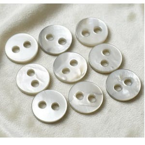 May include: A collection of nine round, off-white buttons with two holes each. The buttons have a pearlescent sheen and are arranged on a light-colored, textured surface. These buttons are suitable for sewing and craft projects.