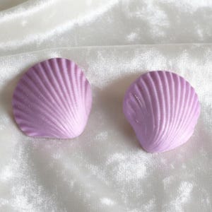 May include: Two lavender-colored seashell-shaped objects with a ribbed texture. The shells are arranged on a white, textured surface. The shells have a gradient effect, fading from lavender to white.