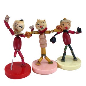 May include: Three vintage-style figurines crafted from wooden beads and pipe cleaners. Each figure features a painted face, articulated limbs, and stands on a circular base. The figures are colored in red, pink, and cream, with varied bead shapes.