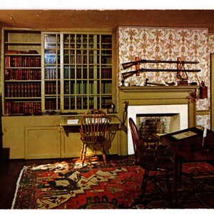 May include: Interior shot of a room with a library, fireplace, and antique furniture. Bookshelves are filled with books. Two rifles are mounted above the fireplace. A patterned rug covers the floor, and sunlight streams in from a window.