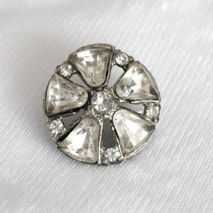 May include: A silver-toned button with a floral design. The button features large, clear, faceted crystals in a circular pattern, with smaller crystals between them. The center has a single, smaller crystal.