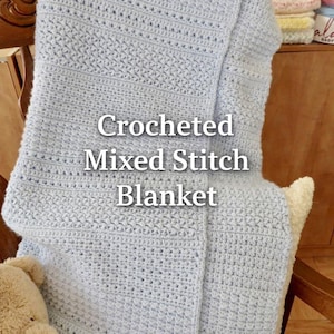 May include: A light blue crocheted blanket with a mixed stitch pattern. The blanket is folded over a wooden chair and features the text "Crocheted Mixed Stitch Blanket". A teddy bear is visible in the lower left corner.
