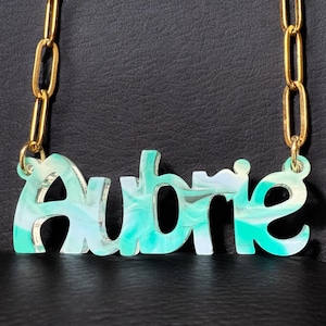 May include: A gold chain necklace with a turquoise and white marbled acrylic nameplate that says "Aubrie".