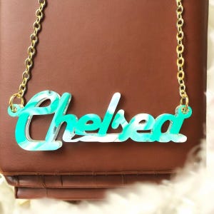 May include: A gold chain necklace with a pendant spelling "Chelsea" in a teal and white marbled design. The pendant is made of acrylic and is suspended from a delicate gold chain. The necklace is displayed on a brown surface.
