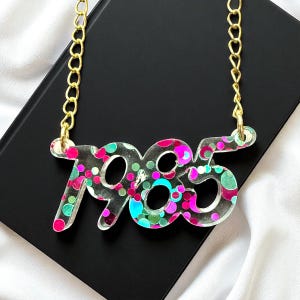 May include: A clear acrylic necklace with the number "1985" in a bold font, filled with pink, green, and blue glitter. The pendant hangs from a gold-colored chain. The necklace is displayed on a black surface.