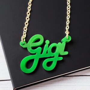 May include: A bright green acrylic necklace spelling "Gigi" in a cursive font. The pendant hangs from a delicate gold-colored chain. The necklace is displayed on a black surface.