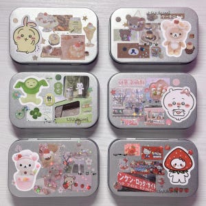 Könnte beinhalten: Sechs kleine, rechteckige, silberfarbene Dosen, die mit Aufklebern verziert sind. Jede Dose hat ein anderes Design, darunter Cartoon-Figuren, Desserts und japanischer Text. Die Dosen sind in einem Raster angeordnet und zeigen eine Vielzahl farbenfroher und skurriler Themen.
