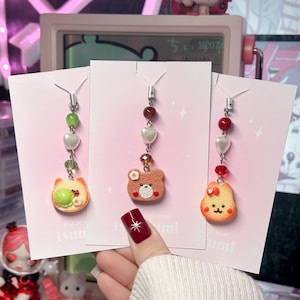 Pode incluir: Três pares de brincos pendurados em cartões rosa. Cada brinco apresenta um pingente com um design diferente: um gato, um urso e uma fatia de pão sorrindo. Os brincos têm contas decorativas e detalhes em forma de coração. Os cartões têm o texto "tsumi" e "with love".