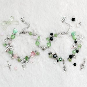 Puede incluir: Dos pulseras de cuentas con dijes. Una pulsera presenta cuentas rosas y verdes, lazos plateados y un dije de corazón. La otra pulsera tiene cuentas negras y verdes, un dije de estrella y un dije de guitarra. Hay dijes y cuentas adicionales dispersos.